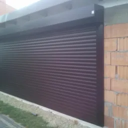 Zamknięta, brązowa roleta zewnętrzna na garażu z ceglaną ścianą w tle. Widoczne elementy konstrukcyjne i trawa.