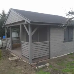 Szara altana z drewnianą podłogą i balustradą, widok z zewnątrz. Elementy konstrukcyjne pomalowane na szaro, dach pokryty ciemną dachówką.