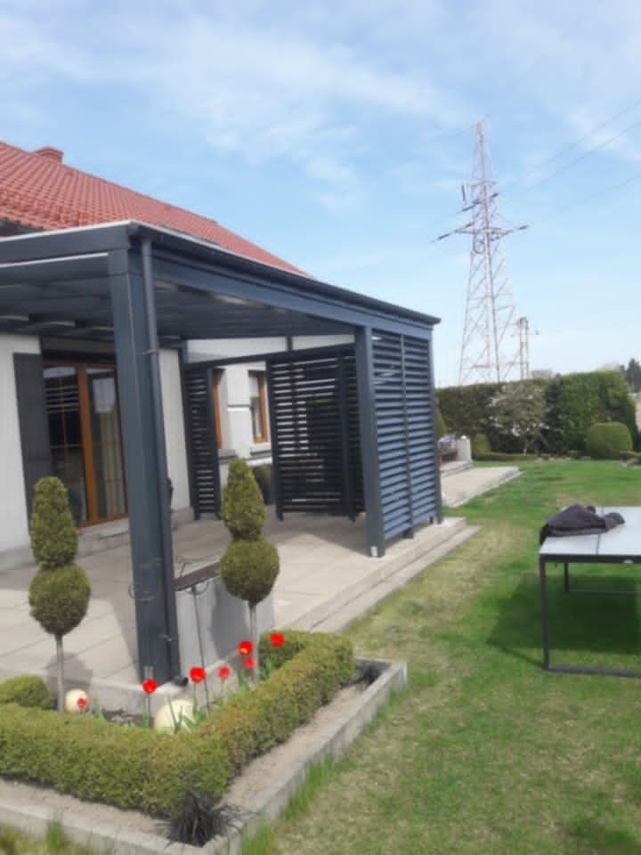 Nowoczesna pergola z szarymi lamelami, przylegająca do białego domu z czerwoną dachówką. Uwagę przyciągają formowane krzewy i rabata z czerwonymi tulipanami.