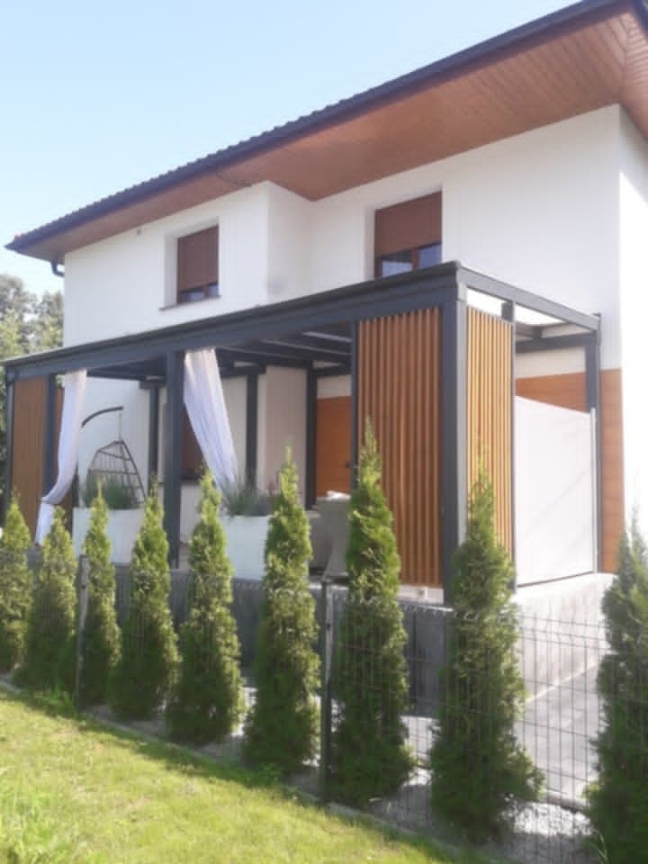 Nowoczesna pergola z drewnianymi panelami i białymi zasłonami przy białym domu, otoczona tujami i metalowym ogrodzeniem.