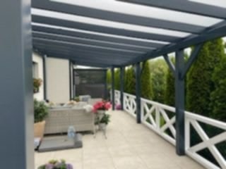 Zadaszony taras z drewnianą pergolą w kolorze antracytowym, biała balustrada z krzyżowym wzorem, jasna podłoga i meble ogrodowe. Widok na zieleń.