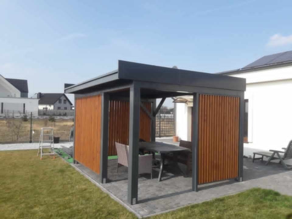 Nowoczesna altana z drewnianymi panelami i szarą konstrukcją, stół z krzesłami w środku, kostka brukowa na podłożu, trawnik i dom w tle.