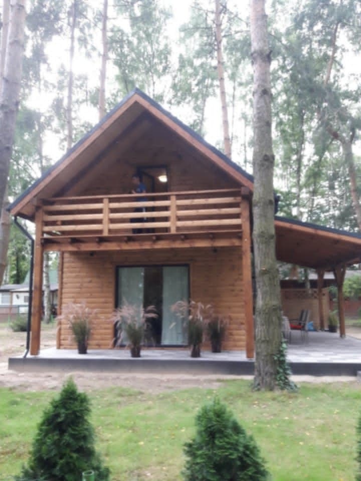 Dwukondygnacyjny dom szkieletowy z drewnianym balkonem i zadaszonym tarasem, otoczony drzewami. Na balkonie stoi osoba. Przed wejściem ozdobne trawy w donicach.