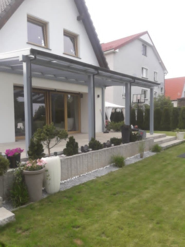 Nowoczesna pergola z szarymi słupami, przylegająca do białego domu, z widokiem na zadbany ogród i taras. Elementy dekoracyjne: donice z kwiatami.