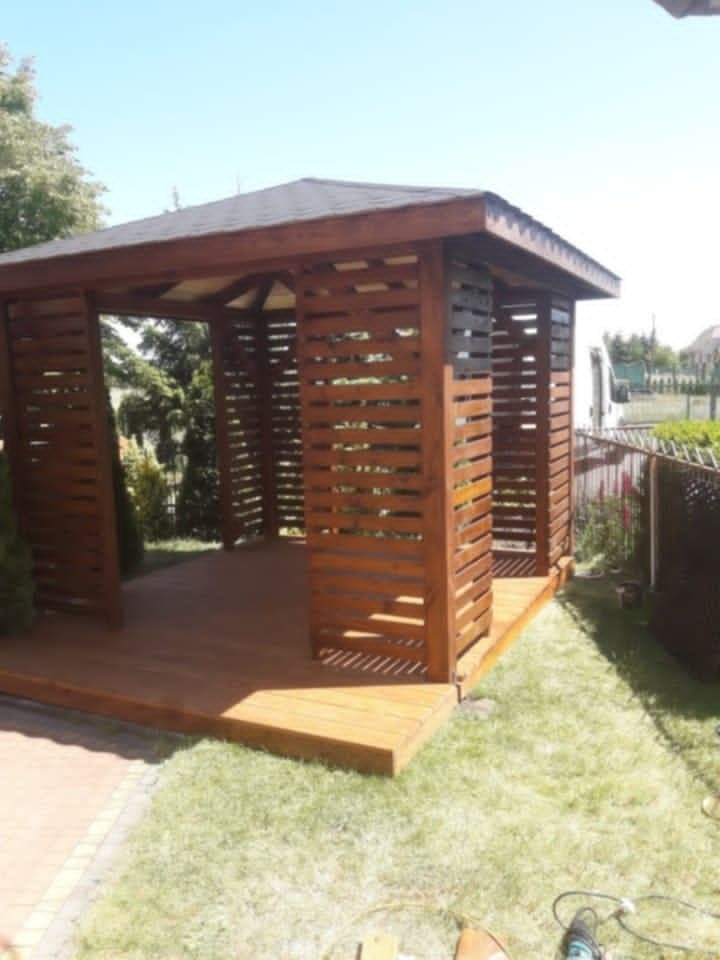 Drewniana altana z ażurowymi ścianami i ciemnym dachem, osadzona na drewnianym podeście, otoczona zielenią. Widoczne narzędzia.