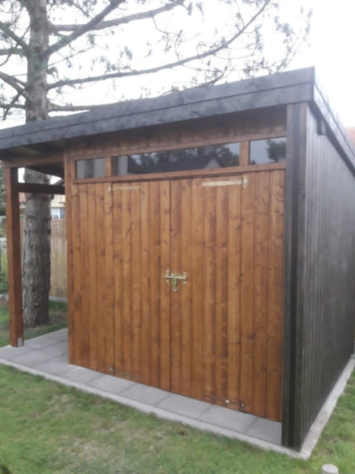 Funkcjonalna, drewniana altanka z dwuskrzydłowymi drzwiami i poziomym oknem, osadzona na betonowej podmurówce, otoczona zielonym trawnikiem i drzewem.