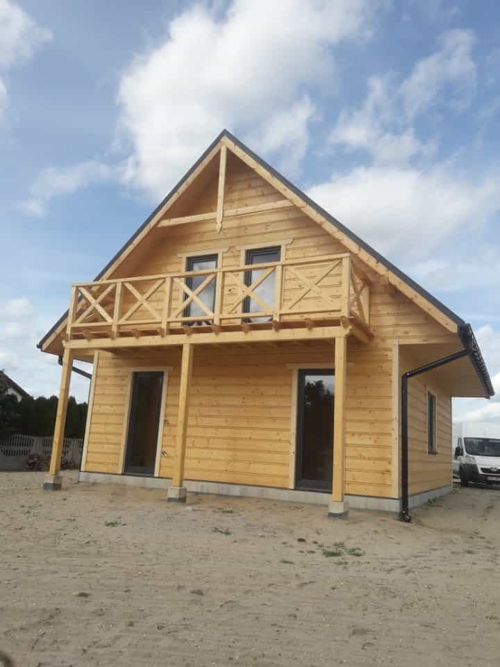 Surowy, dwukondygnacyjny dom szkieletowy z drewnianym balkonem i czarną stolarką okienną, na tle błękitnego nieba z chmurami.