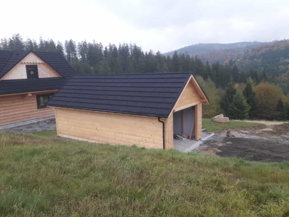 Surowy, drewniany garaż z czarnym dachem w Ostrzeszowie, obok dom z bali na tle lasu i wzgórz. Widok z bliska, pochmurna pogoda.