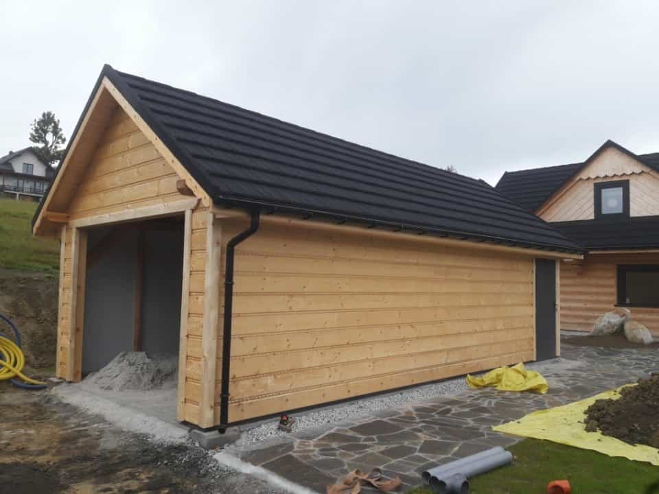 Drewniany garaż z czarnym dachem i otwartym wjazdem, obok domu w stylu góralskim, na tle pochmurnego nieba. Widoczne materiały budowlane.