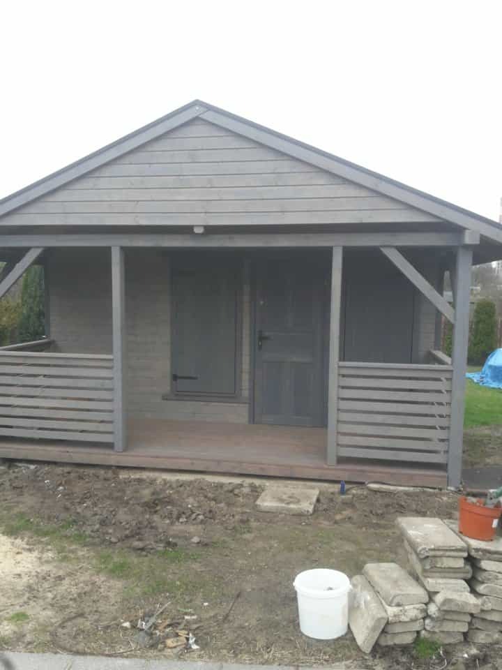 Szara altana z drewnianą podłogą i barierkami, widok z zewnątrz, z elementami ogrodu w tle. Widoczne materiały budowlane i doniczka.