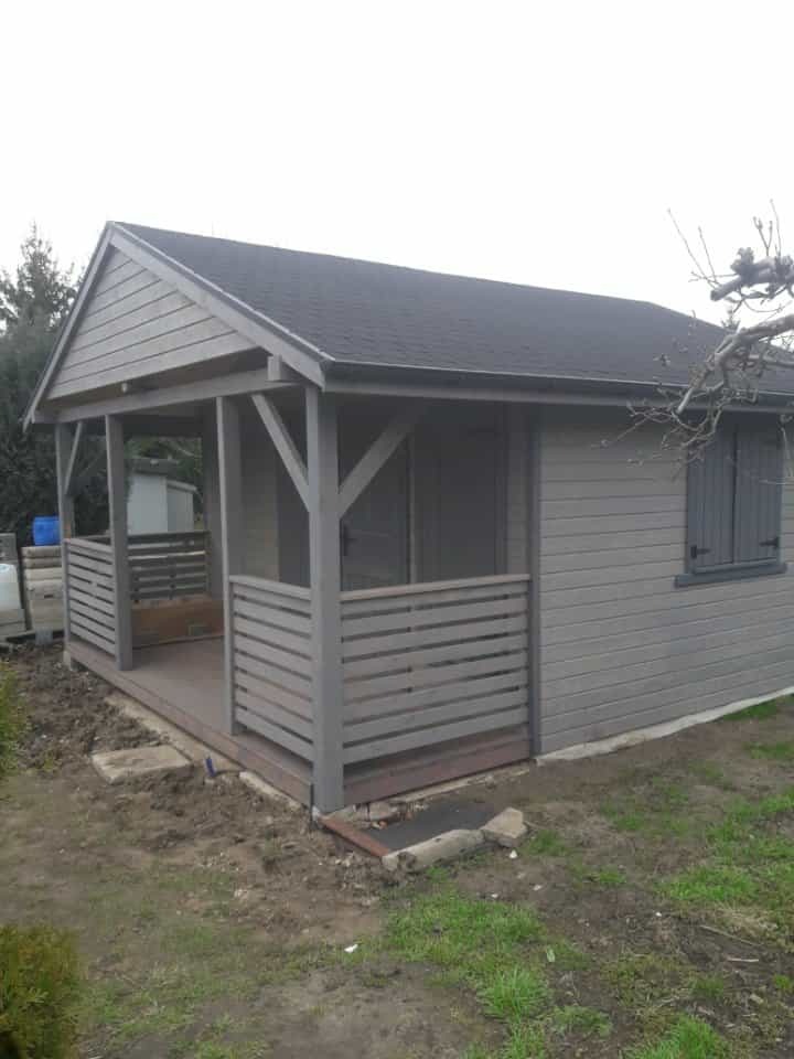 Szara altana z drewnianą podłogą i balustradą, widok z zewnątrz. Elementy konstrukcyjne pomalowane na szaro, dach pokryty ciemną dachówką.