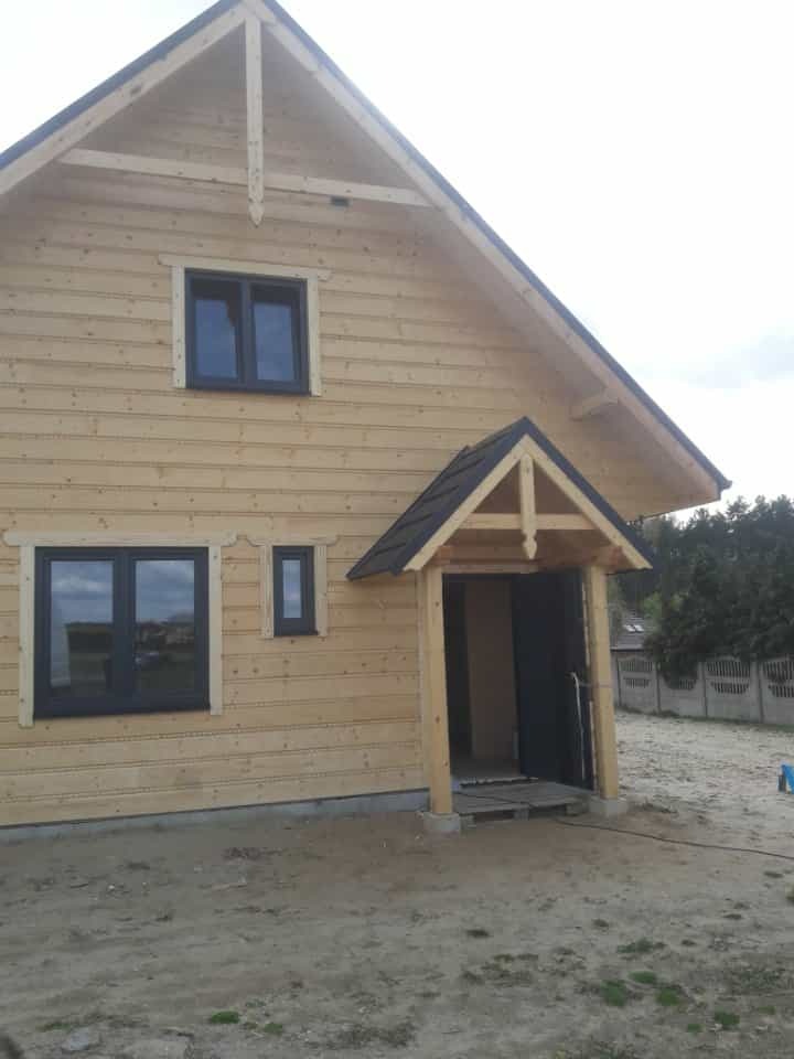 Surowy dom szkieletowy z jasnego drewna, z ciemnymi oknami i zadaszonym wejściem. Widoczne niedokończone otoczenie.