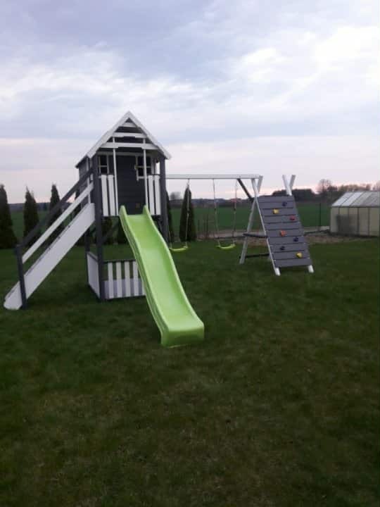 Kompleksowy plac zabaw z domkiem, zjeżdżalnią i ścianką wspinaczkową na trawiastym terenie. Widoczne huśtawki. Całość utrzymana w szaro-białej kolorystyce.