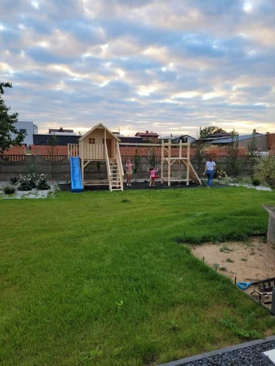 Drewniany plac zabaw z domkiem, zjeżdżalnią i huśtawką na trawiastym podwórku. W tle ogrodzenie i budynki mieszkalne, a na pierwszym planie rozległy trawnik.