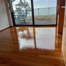 Błyszcząca, nowo położona drewniana podłoga z widokiem na morze i drzewo przez duże okno balkonowe. Odbicia w podłodze podkreślają jej gładkość i połysk.