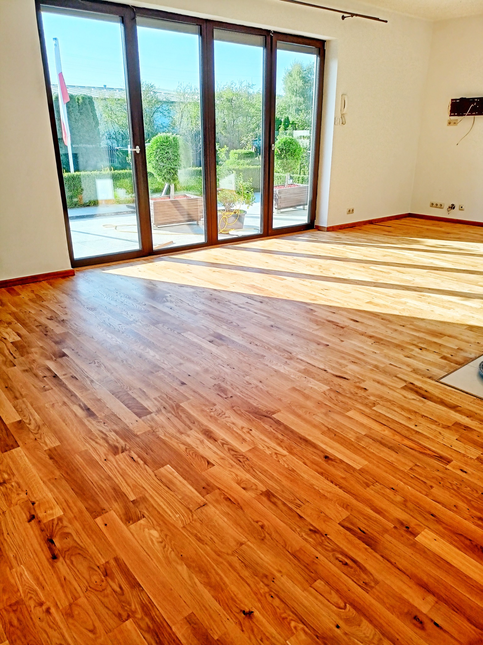 Jasne, lakierowane deski parkietowe w nowym wnętrzu, z widocznymi promieniami słonecznymi wpadającymi przez duże okna tarasowe.