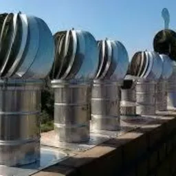 Rząd metalowych nasad kominowych obrotowych na dachu budynku, widoczne otoczenie z drzewami i budynkami w oddali, błękitne niebo.