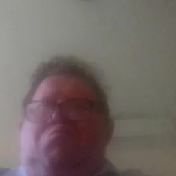 Rozmazane zdjęcie twarzy mężczyzny w okularach na tle jasnej ściany.