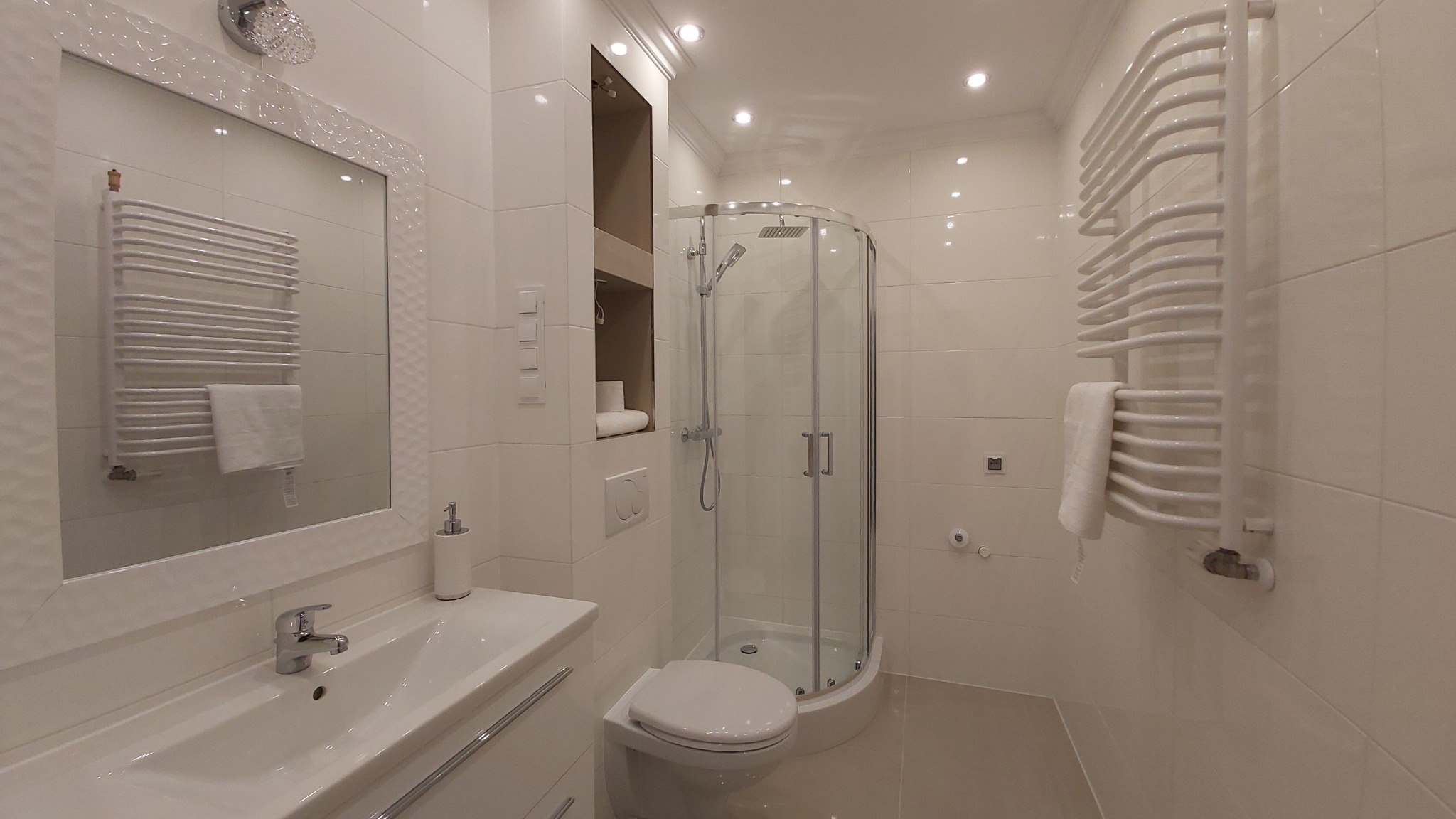 Jasna łazienka z kabiną prysznicową narożną, umywalką w zabudowie, grzejnikiem drabinkowym z ręcznikiem oraz wnęką z półkami.