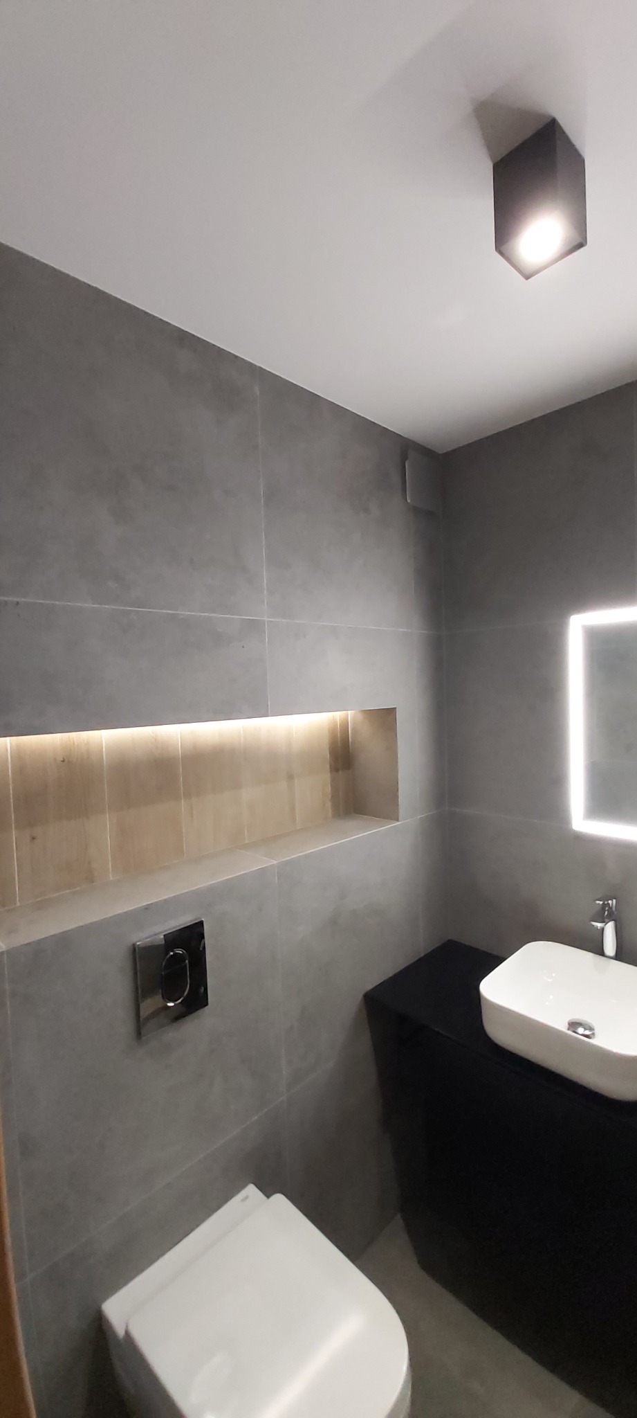 Nowoczesna łazienka z szarymi płytkami imitującymi beton, podświetloną wnęką, kwadratowym lustrem z podświetleniem LED, wiszącą toaletą i czarną szafką pod umywalką.