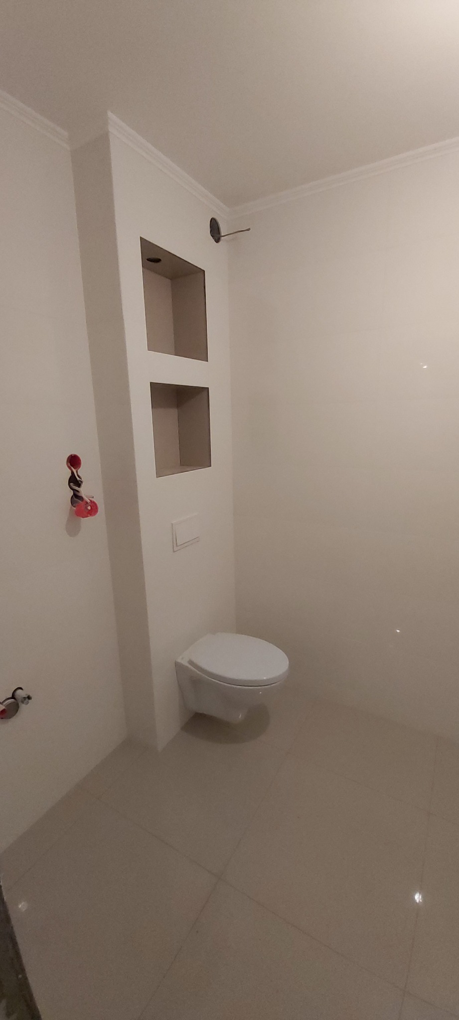 Wykończona łazienka z podwieszaną toaletą, zabudową z półkami i białymi płytkami na ścianach i podłodze, widoczne przyłącza hydrauliczne.