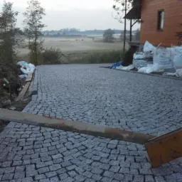 Nowo ułożona nawierzchnia z kostki w odcieniach szarości w kształcie wachlarza, z widocznymi workami z materiałem budowlanym przy drewnianym domu w tle, w porannym świetle.