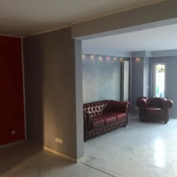 Salon po odświeżeniu z bordową skórzaną sofą typu Chesterfield i marmurową podłogą, widok z przyległego pokoju z czerwoną ścianą.