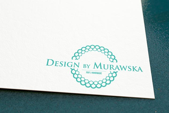 Logo 'Design by Murawska' w turkusowym kolorze, otoczone dekoracyjnym, okrągłym wzorem, wydrukowane na kremowym papierze o delikatnej fakturze.