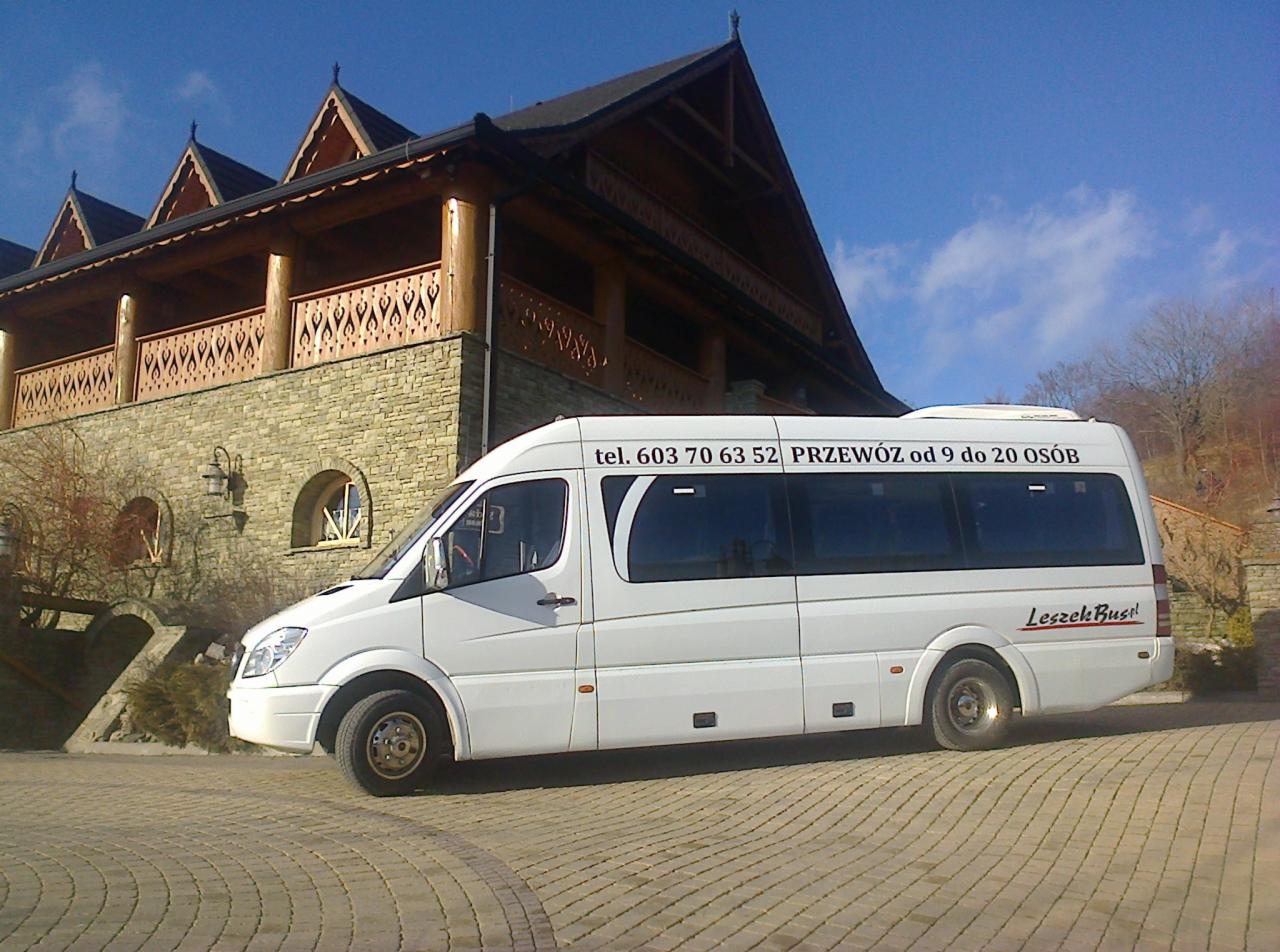 Biały bus z napisem 'Leszek Bus' i numerem telefonu na tle drewnianego budynku w stylu góralskim, na brukowanym podjeździe.