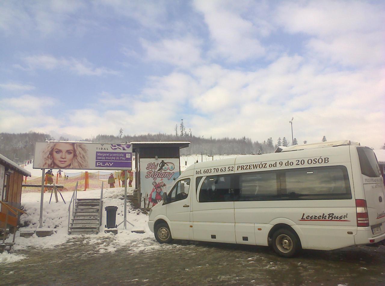 Biały bus firmy Leszek Bus.pl z napisem 'Przewóz od 9 do 20 osób' zaparkowany na ośnieżonym parkingu przy stoku narciarskim z reklamą Tidal i Ski Siglany w tle.