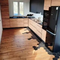 Nowoczesna kuchnia z drewnopodobnymi frontami szafek i czarnymi elementami, drewniana podłoga przechodząca w heksagonalne płytki, czarny sprzęt AGD.