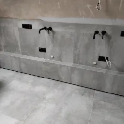 Surowy, minimalistyczny wystrój łazienki w trakcie wykańczania: szare płytki wielkoformatowe na ścianach i podłodze, czarne baterie umywalkowe montowane na ścianie, widoczne niedoskonałości...