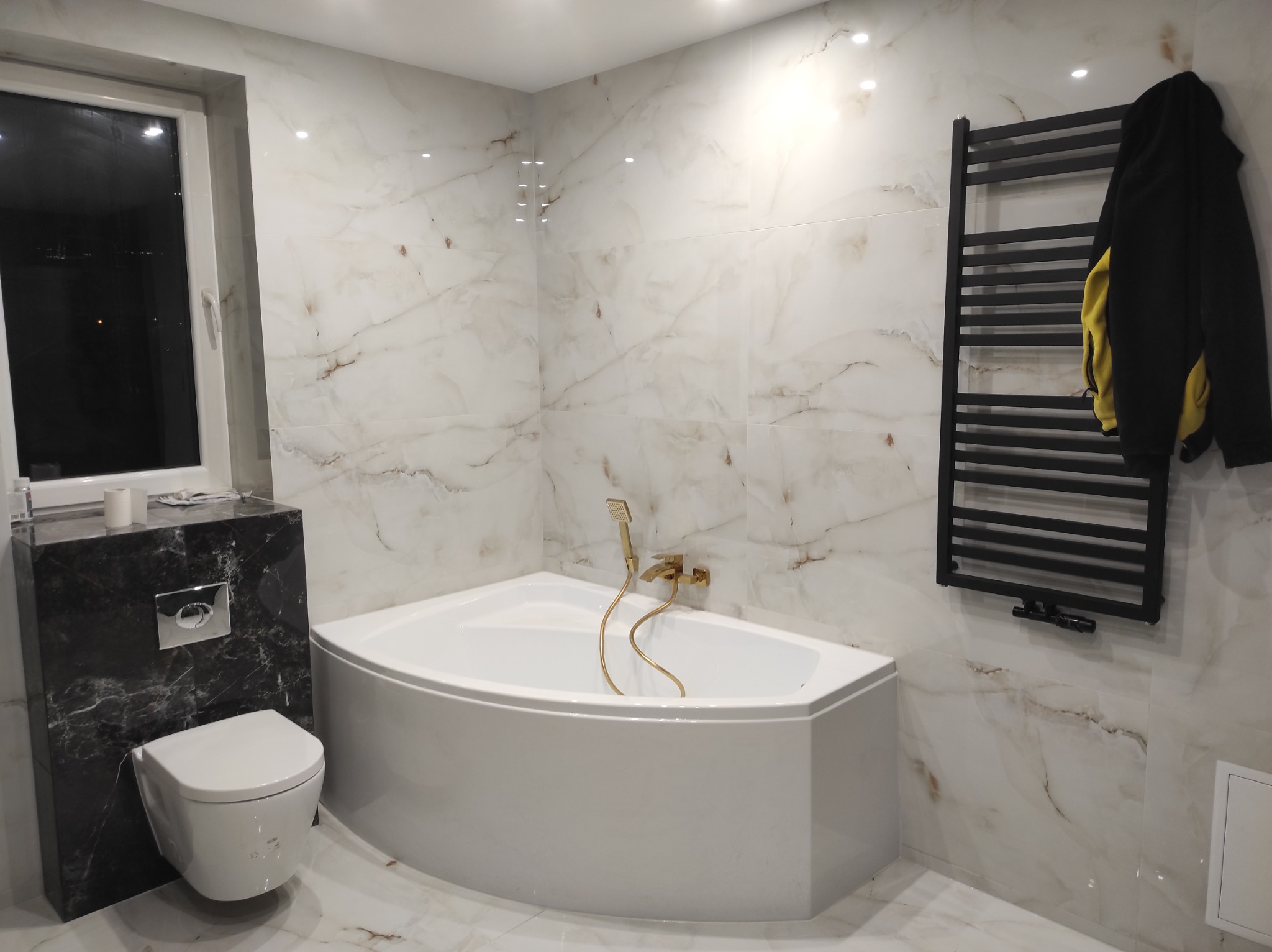 Nowoczesna łazienka z narożną wanną, marmurowymi płytkami, podwieszaną toaletą i czarnym grzejnikiem drabinkowym z wiszącą bluzą.