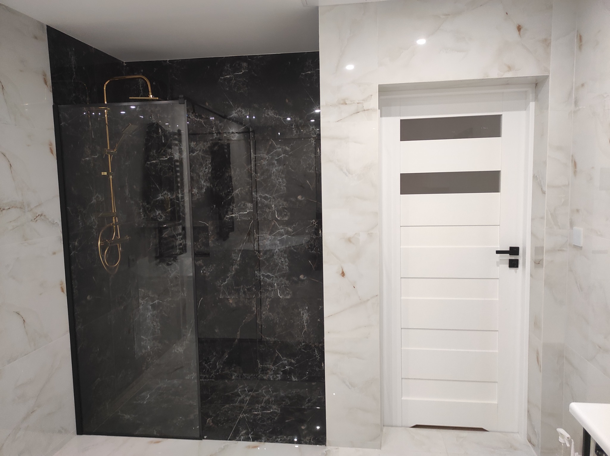 Wykończona łazienka z kabiną prysznicową typu walk-in z czarnymi marmurkowymi płytkami, złotą armaturą i białą, nowoczesną stolarką drzwiową na tle jasnych, marmurkowych płytek.