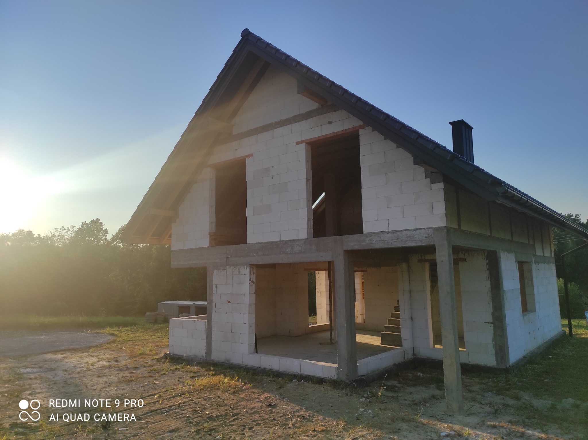 Surowy stan domu jednorodzinnego z białego bloczka, widoczne otwory okienne i drzwiowe, dach pokryty ciemną dachówką, w tle zieleń drzew, wczesne poranne słońce.