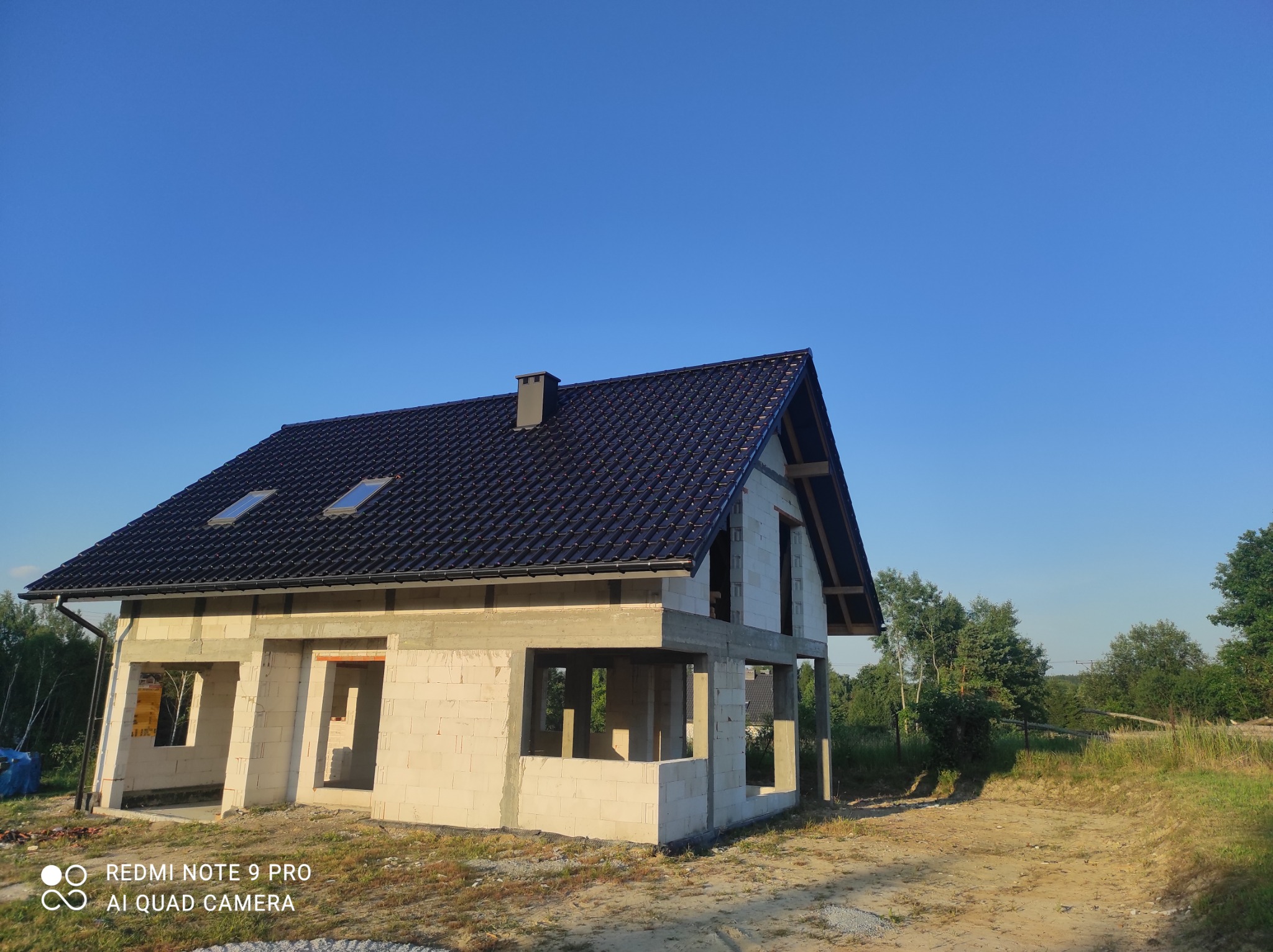 Dom murowany w stanie surowym zamkniętym z ciemną dachówką i oknami dachowymi, na tle błękitnego nieba i zieleni drzew.