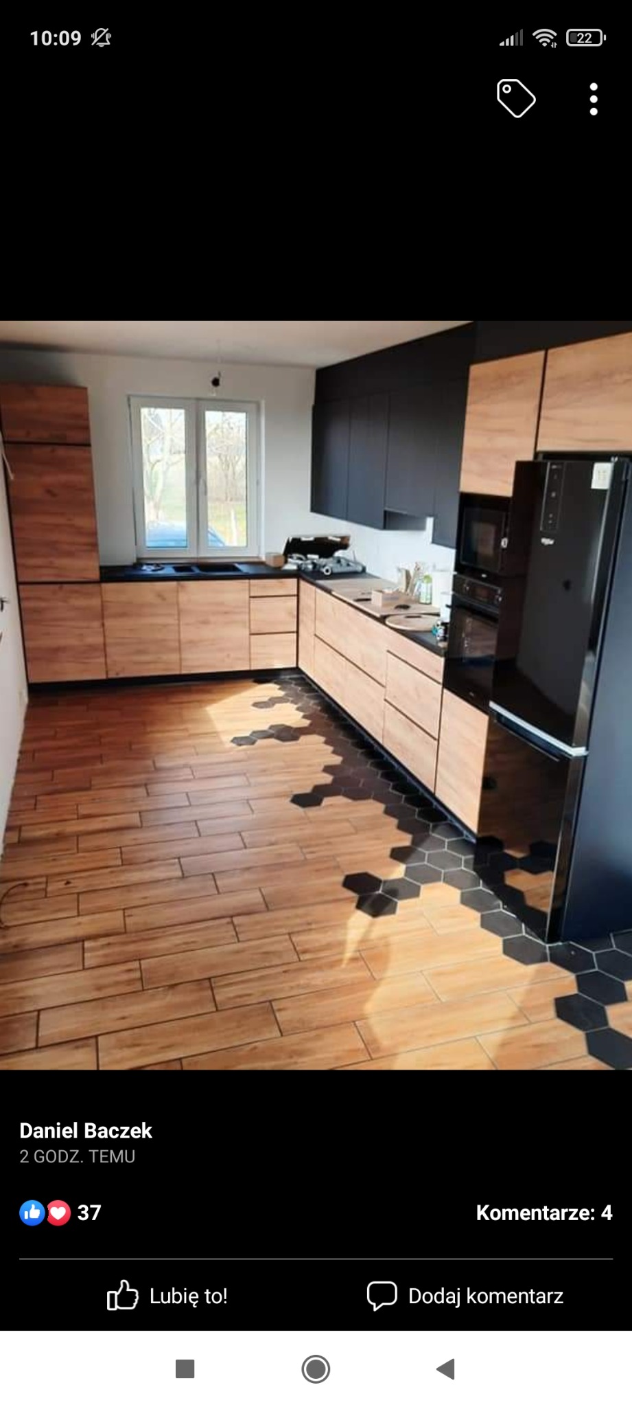 Nowoczesna kuchnia z drewnopodobnymi frontami szafek i czarnymi elementami, drewniana podłoga przechodząca w heksagonalne płytki, czarny sprzęt AGD.