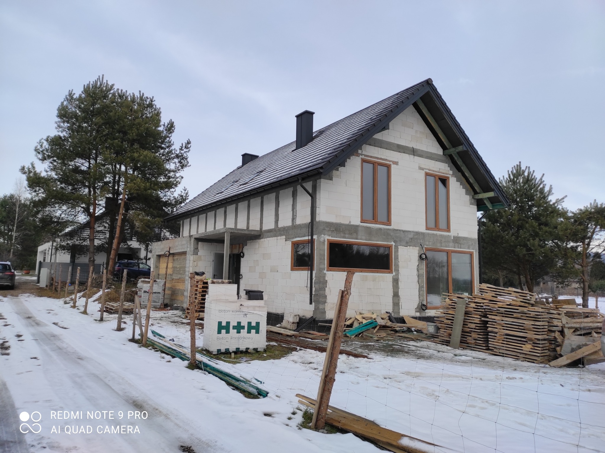 Niedokończony dom murowany z bloczków H+H w zimowej scenerii, widoczne drewniane okna, czarny dach i kominy, oraz materiały budowlane na posesji.