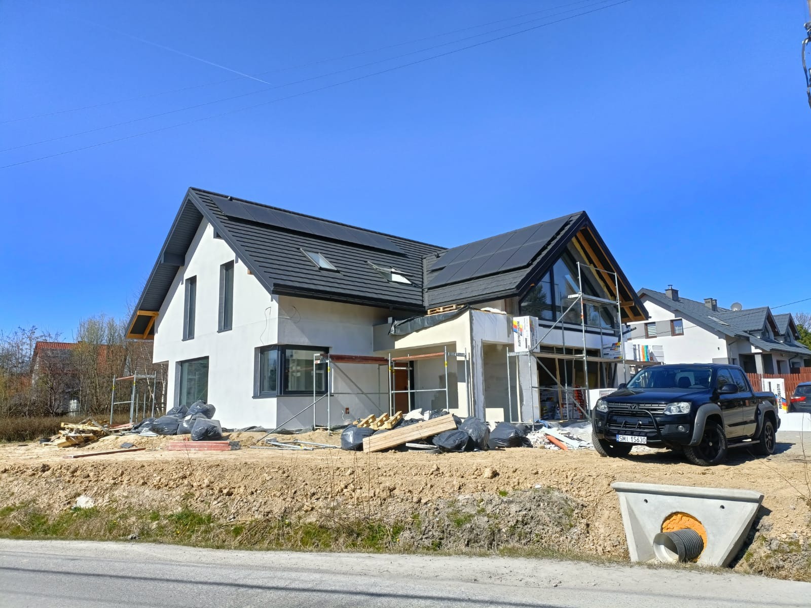 Budowa domu jednorodzinnego z czarną dachówką i dużymi oknami, widoczne rusztowania i czarny pickup na tle błękitnego nieba.