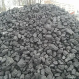 Góra czarnego, błyszczącego węgla kamiennego ułożona na zewnątrz, z widocznymi fragmentami samochodów i budynków w tle.