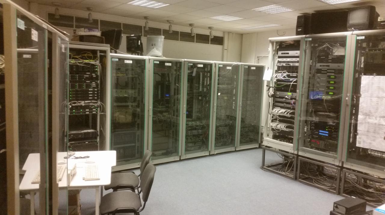 Rząd szaf serwerowych w klimatyzowanym pomieszczeniu data center, widoczne kable i urządzenia sieciowe, na pierwszym planie biurko z klawiaturami i okularami.