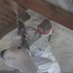 Pracownik w kombinezonie ochronnym aplikuje izolację natryskową na poddaszu, widoczne drewniane elementy konstrukcyjne dachu.