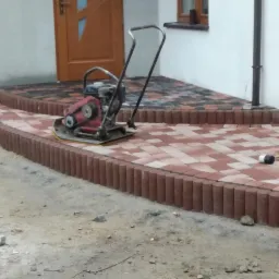 Czerwono-szara kostka ułożona na podjeździe z użyciem zagęszczarki w pobliżu wejścia do budynku.