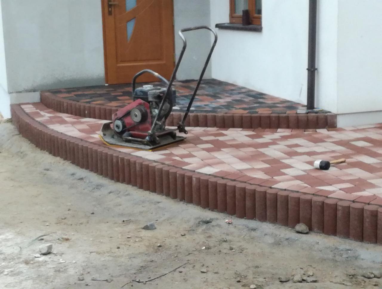 Czerwono-szara kostka ułożona na podjeździe z użyciem zagęszczarki w pobliżu wejścia do budynku.