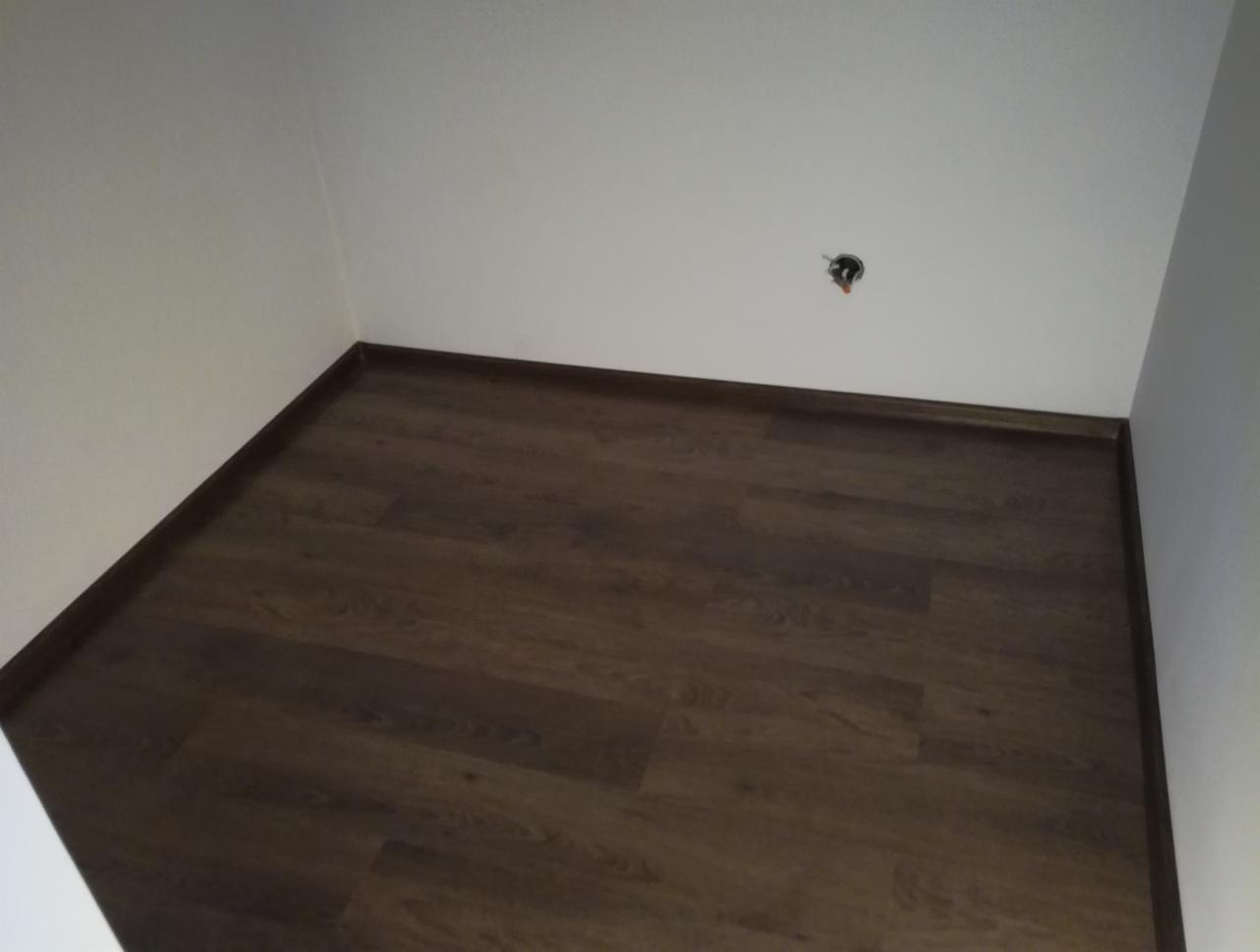 Ukończona podłoga z paneli imitujących ciemne drewno, z widocznymi listwami przypodłogowymi i gniazdkiem elektrycznym w ścianie.