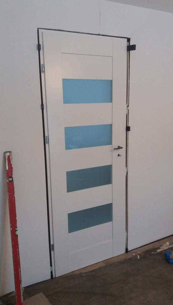 Białe drzwi wewnętrzne z czterema poziomymi, błękitnymi przeszkleniami, zamontowane w surowym otworze, z widoczną poziomicą i narzędziami na podłodze.