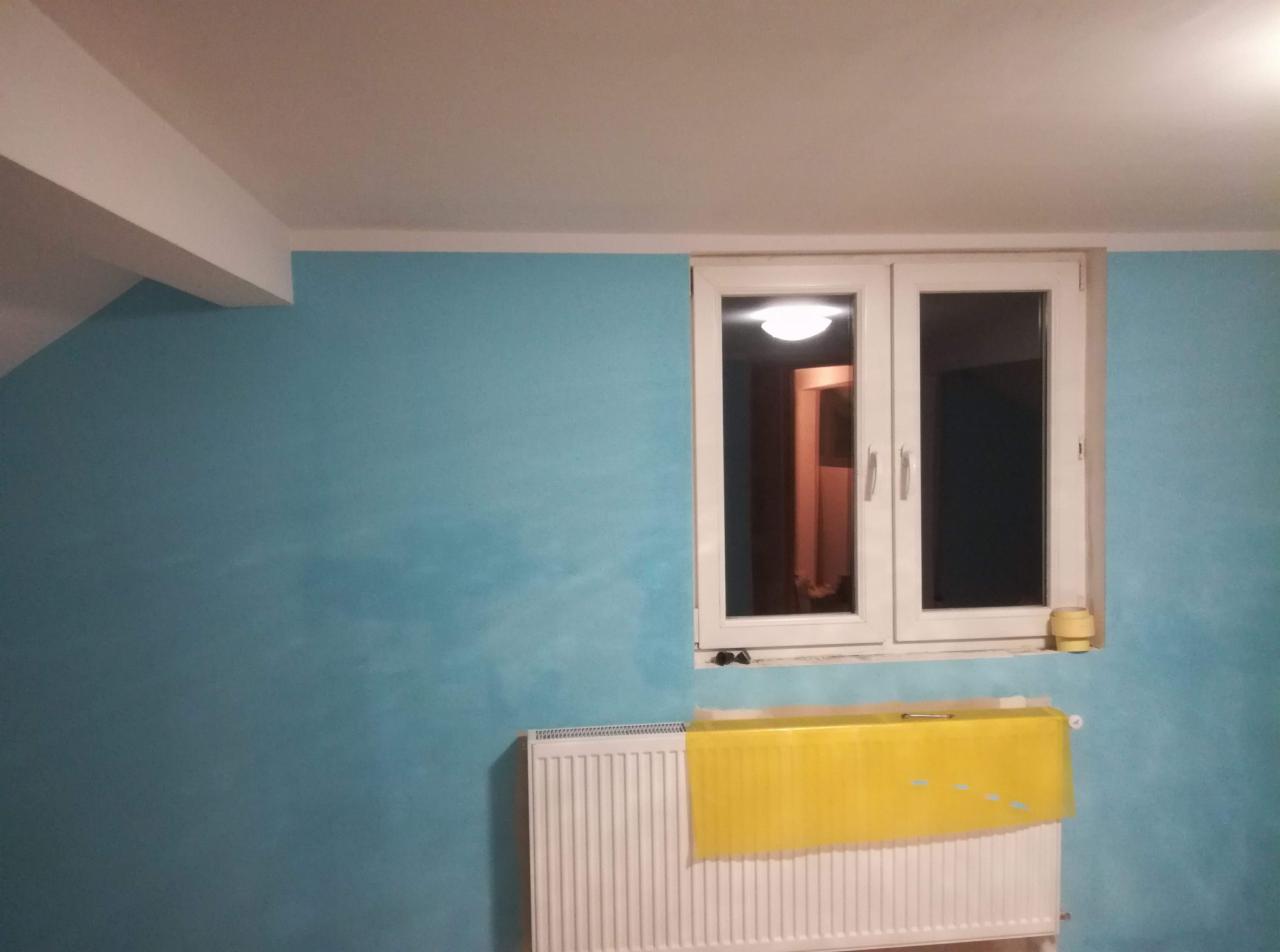 Błękitna ściana po malowaniu z widocznym grzejnikiem zabezpieczonym żółtą folią malarską i oknem z widokiem na wnętrze.