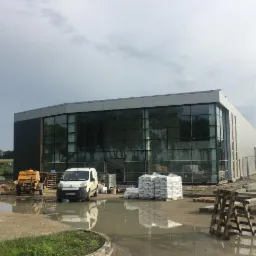 Nowoczesny budynek w trakcie budowy z dużymi oknami, maszyna Putzmeister i biały van na placu budowy, worki z materiałami budowlanymi, kałuże po deszczu.