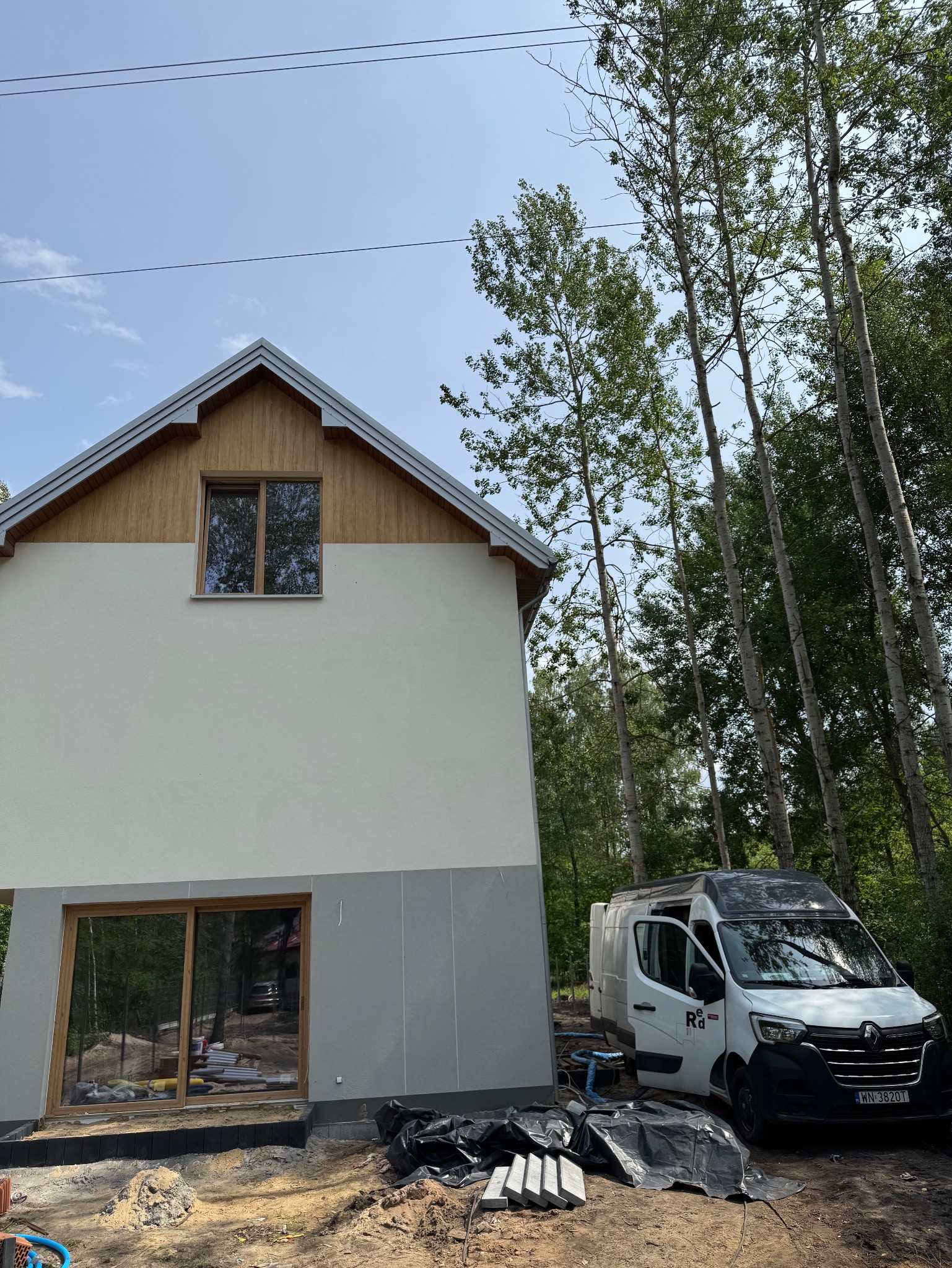 Nowoczesny dom z oknami z drewna, świeżo otynkowaną elewacją w odcieniach bieli i szarości. Obok zaparkowany biały bus firmy budowlanej, wysokie drzewa w tle.