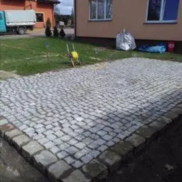 Ułożona nawierzchnia z kostki granitowej w szarym odcieniu, obrzeża z większych kamieni, trawnik, budynek mieszkalny i zaparkowany samoch&oacute;d dostawczy w tle.